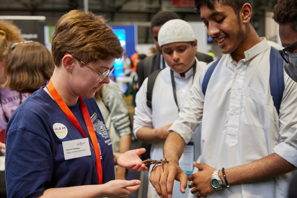 Claire Hoarau from Harper Adams University gudes handling of a New Guinea spiny stick insect by two boys, on the Royal Entomological Society stand at The Big Bang Fair 2022